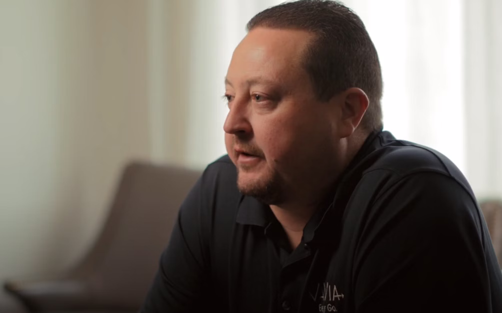 A man in a black shirt sits comfortably in a chair, appearing thoughtful and attentive to his environment.